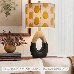 May include: A decorative table lamp with a beige and black base and a cylindrical lampshade. The lampshade features a pattern of yellow circles on a beige background. The base has an oval cutout. The text at the bottom says "STYLED FOR INSPIRATION - LISTING IS FOR LAMPSHADE ONLY."