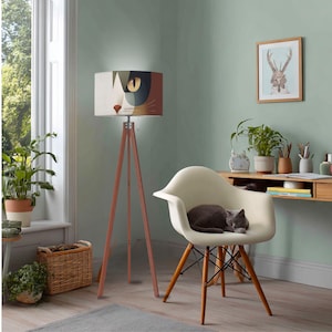 May include: A floor lamp with a tripod wooden base and a cylindrical lampshade featuring a stylized cat face graphic. The lamp stands next to a cream-colored molded armchair with a gray cat resting on the seat cushion.