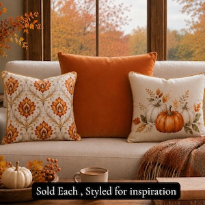 May include: Three square throw pillows on a sofa. The left pillow features an orange and gold floral damask pattern. The center pillow is solid burnt orange. The right pillow displays a printed graphic of pumpkins and autumn leaves. Text reads: Sold Each , Styled for inspiration.