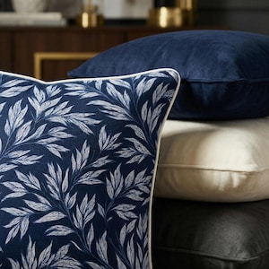 May include: A stack of decorative throw pillows in various colors and textures. The front pillow features a navy blue background with a white leaf pattern and white trim. Other pillows are navy blue, cream, and black.