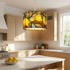 May include: A cylindrical pendant light with a stained glass lampshade. The lampshade has a design of yellow lemons, green leaves, and brown branches. The light hangs from a white cord in a kitchen setting.