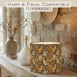 May include: A decorative lamp with a white ceramic base and a gold-toned harp. The lampshade features a pattern of birds and foliage in shades of gold, brown, and green. The text at the top reads "Harp & Finial Compatible (Upgrade)".