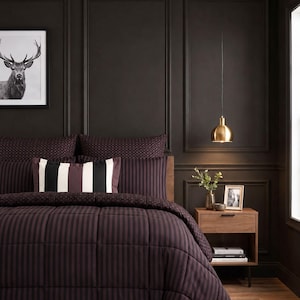 May include: A bedding set featuring a dark purple and black striped comforter and matching pillow shams. A rectangular accent pillow with vertical black, white, and purple stripes rests against the pillows. The bedding is set against a dark wall.