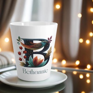 Peut inclure: Tasse à café en céramique blanche avec un motif décoratif. Le motif représente un oiseau bleu, la lettre "B" et le nom "Bethannie". L'oiseau a des plumes orange et marron. La tasse est posée sur une soucoupe blanche.