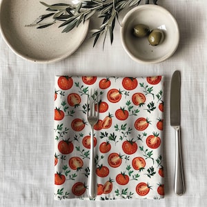 May include: A white cloth napkin with a repeating pattern of red tomatoes and green sprigs. A silver fork and knife are placed on the napkin. A beige plate and a small bowl with olives are also visible.