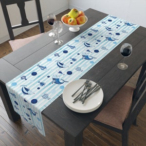 May include: A blue and white table runner with a striped pattern and a watercolor design of fish and bubbles. The runner is on a dark wood table with a white plate and silverware.