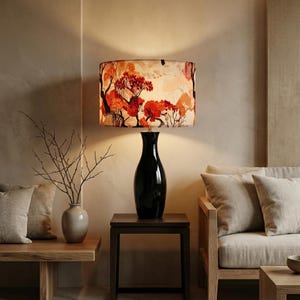 May include: A black table lamp with a patterned lampshade featuring a design of red and orange trees. The lamp sits on a dark brown side table. The room has a neutral color palette with a vase and a sofa.