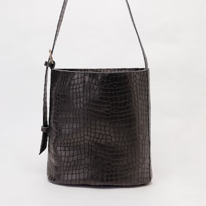 May include: A black bucket bag featuring a textured crocodile skin pattern. The bag has a long, adjustable shoulder strap with a metal buckle and a vertical strap detail on the side. The design is structured with a flat base and an open top opening.