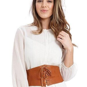 Op de afbeelding: Een witte blouse met lange mouwen en verticale plooien aan de voorzijde, gecombineerd met een brede, lichtbruine korset-stijl riem. De riem is gemaakt van een glad materiaal en heeft een centrale vetersluiting met bijpassende lichtbruine veters.