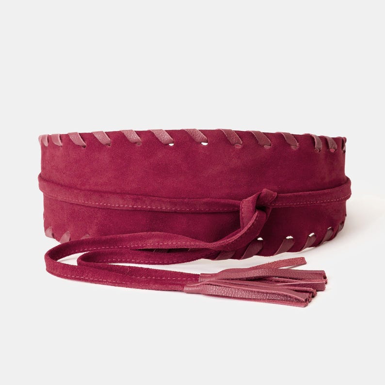 May include: A wide, deep burgundy suede wrap belt featuring a decorative whipstitch edge along the top and bottom borders. The belt includes long, integrated tie straps that finish in matching suede tassels for a secure and adjustable fit.