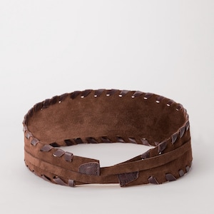 May include: A wide brown suede belt featuring a textured surface and a decorative whipstitch edge detail along the top and bottom borders. The belt is constructed with a wrap-around design and includes a thin integrated strap for secure fastening.