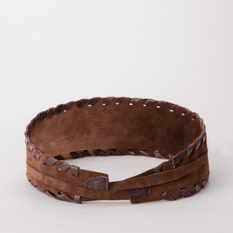 May include: A wide brown suede belt featuring a textured surface and a decorative whipstitch edge detail along the top and bottom borders. The belt is constructed with a wrap-around design and includes a thin integrated strap for secure fastening.
