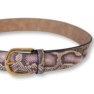 Op de afbeelding: Een riem met een slangenleermotief in roze, crème en donkergrijze tinten. De riem is voorzien van een gepolijste goudkleurige metalen gesp en een bijpassende lus, met een gladde, lichtbeige voering zichtbaar langs de binnenrand.