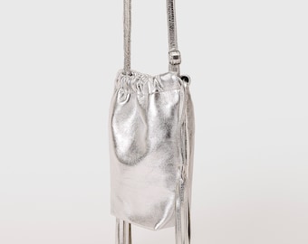 May include: A metallic silver drawstring pouch bag with a smooth, reflective finish. The bag features a long, thin shoulder strap and two decorative tassels hanging from the side. The top is gathered with a drawstring closure, creating a soft, rounded shape.