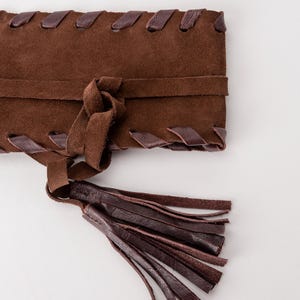 May include: A dark brown suede belt featuring a wide rectangular body with a woven leather lace detail along the top and bottom edges. The belt includes a central tie closure that secures with a knot and ends in a long, multi-strand leather tassel.