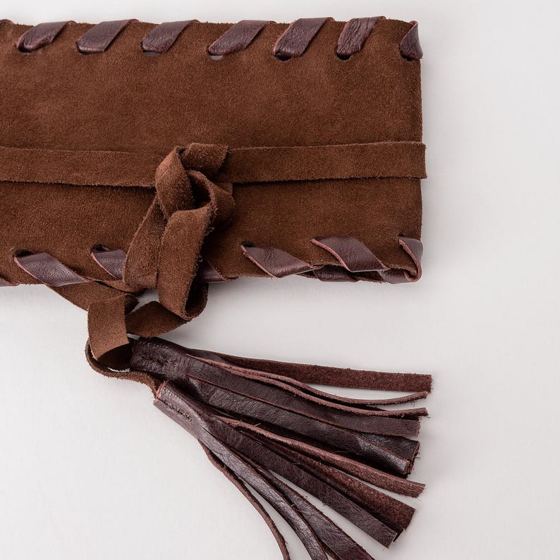 May include: A dark brown suede belt featuring a wide rectangular body with a woven leather lace detail along the top and bottom edges. The belt includes a central tie closure that secures with a knot and ends in a long, multi-strand leather tassel.