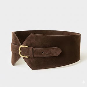 May include: A wide dark brown suede belt featuring a structured, asymmetrical wrap-around design. The belt is secured with a single prong buckle made of brushed brass metal, which is attached to a narrow adjustable strap overlaying the main body.