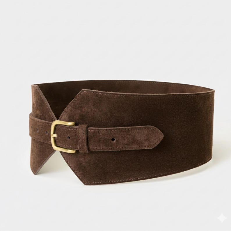 May include: A wide dark brown suede belt featuring a structured, asymmetrical wrap-around design. The belt is secured with a single prong buckle made of brushed brass metal, which is attached to a narrow adjustable strap overlaying the main body.