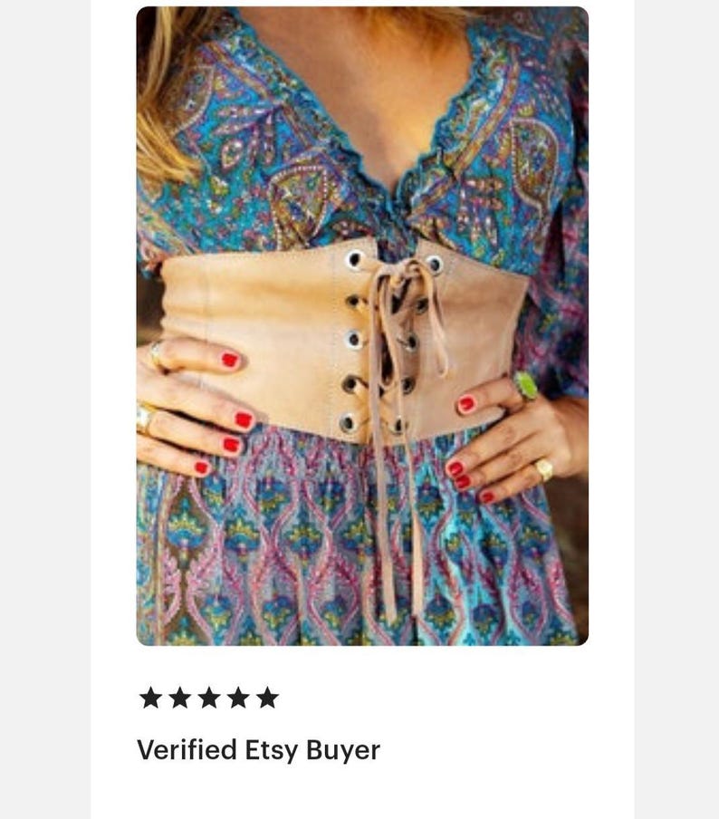 Op de afbeelding: Een brede, beige korset-stijl riem met een vetersluiting aan de voorzijde, zilverkleurige metalen oogjes en bijpassende beige veters. De riem wordt gedragen over een blauwe, turquoise en roze jurk met een paisley-ge&iuml;nspireerd patroon.