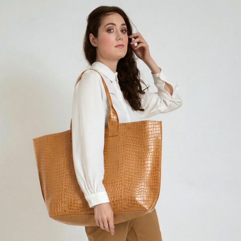 May include: A large tan tote bag featuring a textured crocodile skin pattern. The bag has a rectangular shape with two wide shoulder straps and a spacious main compartment, crafted from a firm material with a consistent embossed finish.