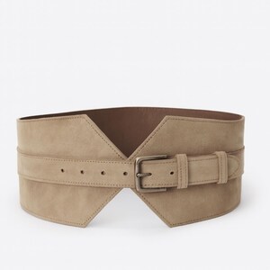 May include: A wide, beige, suede-like corset belt featuring a pointed, V-shaped front design. The belt includes a horizontal strap across the center with a rectangular metal buckle, multiple adjustment holes, and a matching belt loop for securing.
