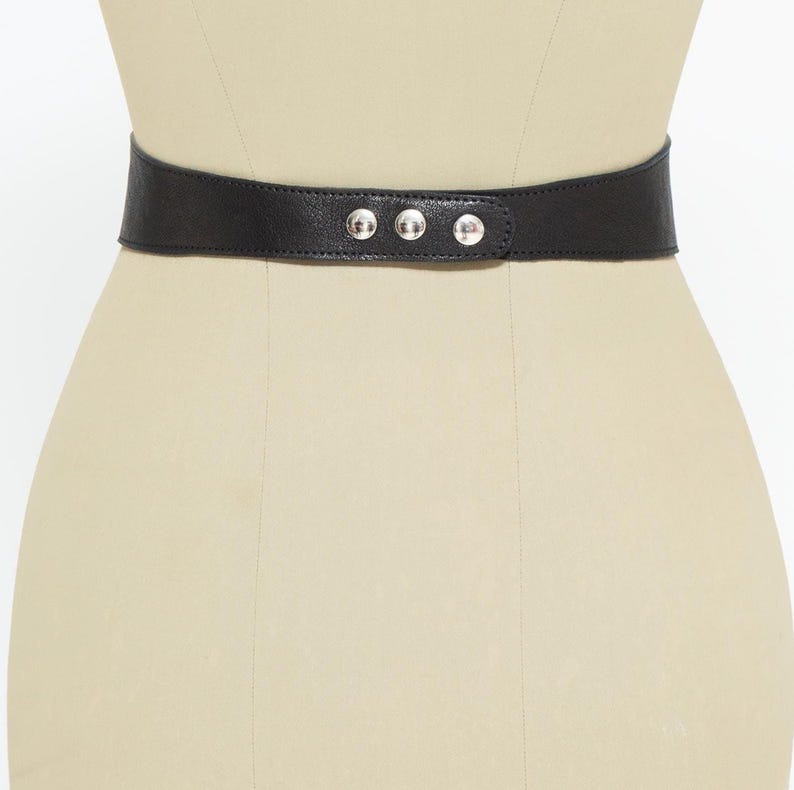 May include: A black leather belt with three silver-colored rivets. The belt is wide and cinches at the waist. The belt is displayed on a mannequin.
