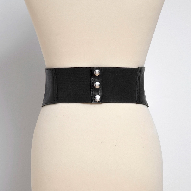 Op de afbeelding: Een brede zwarte elastische tailleband met een centraal verticaal paneel voorzien van drie gepolijste zilverkleurige metalen drukknopen. De riem wordt getoond op een neutraal gekleurde etalagepop, wat de gladde textuur en sluiting benadrukt.
