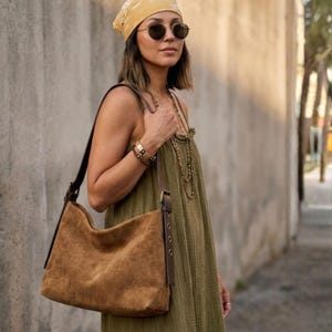 May include: A brown suede shoulder bag with a textured crocodile-embossed pattern, featuring dark brown leather straps with metal buckle hardware, a spacious main compartment, and a slouchy silhouette designed for comfortable over-the-shoulder carrying.