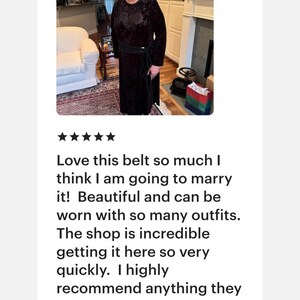 May include: A black belt featuring a textured, patterned surface and a thin, wrap-around tie closure. The belt is worn over a long-sleeved, dark-colored dress with a subtle floral pattern, creating a cinched waist effect with the tie.