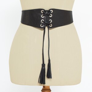 May include: Black corset belt with silver-tone grommets and a lace-up design. The belt has a wide band and two long tassels hanging from the front. The belt is displayed on a mannequin.