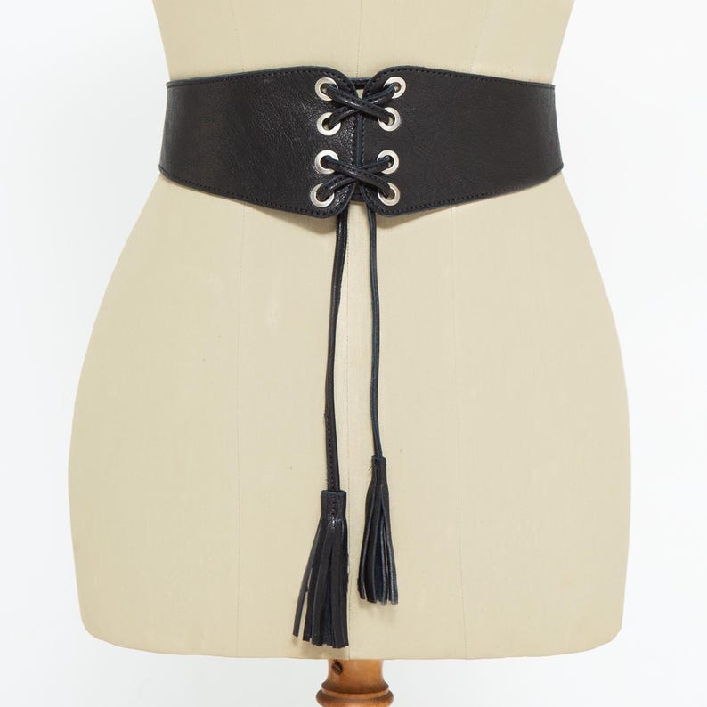 May include: Black corset belt with silver-tone grommets and a lace-up design. The belt has a wide band and two long tassels hanging from the front. The belt is displayed on a mannequin.