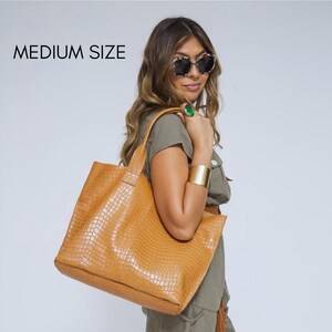 May include: A medium size tan tote bag with a textured crocodile skin pattern. The bag features two shoulder straps and a spacious open-top design. The surface has a glossy finish and a repeating grid-like embossed texture throughout the exterior.