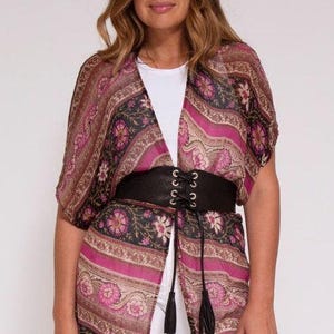 May include: A patterned kimono in shades of pink, brown, and black, worn over a white top. The kimono is cinched at the waist with a black leather belt featuring a lace-up design. The belt has two tassels hanging down.