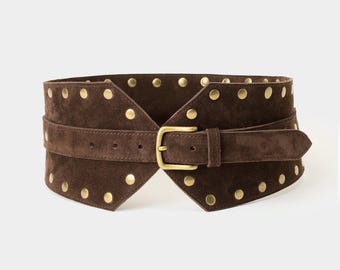 Op de afbeelding: Een brede donkerbruine suède riem met een puntig ontwerp aan de voorzijde, versierd met talloze ronde, messingkleurige metalen studs langs de boven- en onderranden, en vastgezet met een bijpassende messingkleurige metalen gesp.