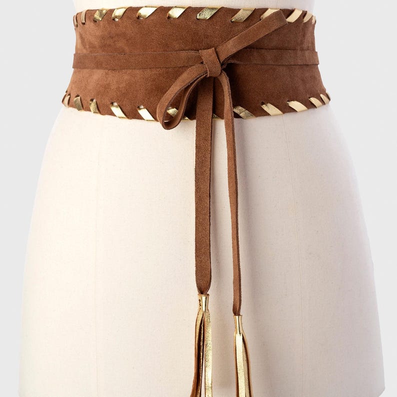 May include: A wide brown suede obi belt featuring a wrap-around tie closure with long dangling ends. The top and bottom edges of the belt are accented with a decorative metallic gold whipstitch lacing pattern, and the tie ends feature gold tassels.