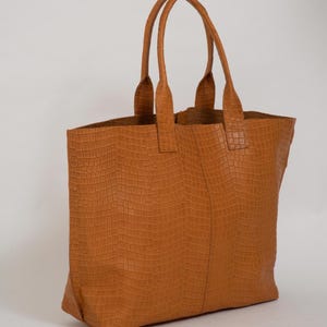 May include: A tan tote bag featuring a textured crocodile-embossed pattern across the entire exterior. The bag has two vertical handles attached to the top edge and a central vertical seam running down the front panel of the rectangular body.