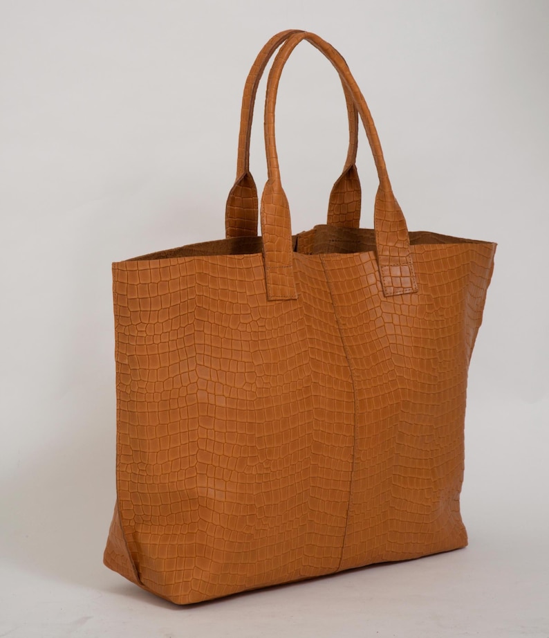 May include: A tan tote bag featuring a textured crocodile-embossed pattern across the entire exterior. The bag has two vertical handles attached to the top edge and a central vertical seam running down the front panel of the rectangular body.