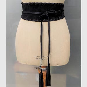 May include: A wide black waist belt featuring a textured suede-like finish, decorative whipstitch detailing along the top and bottom edges, and a thin wrap-around tie closure that secures in the front with two long hanging tassels at the ends.