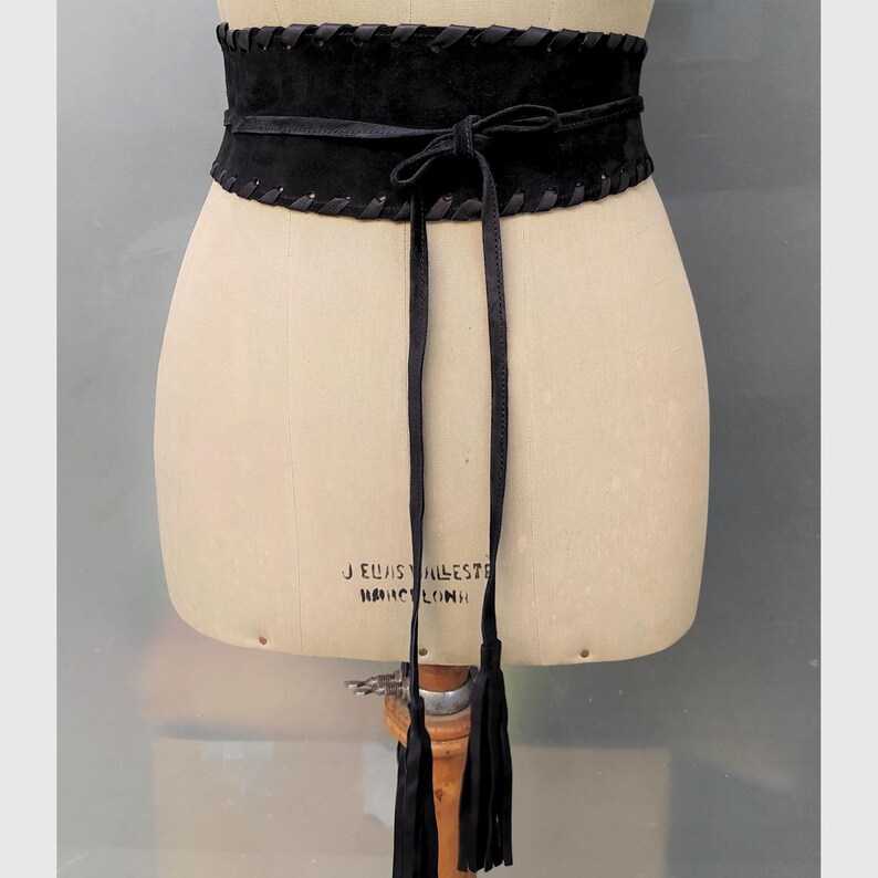 May include: A wide black waist belt featuring a textured suede-like finish, decorative whipstitch detailing along the top and bottom edges, and a thin wrap-around tie closure that secures in the front with two long hanging tassels at the ends.