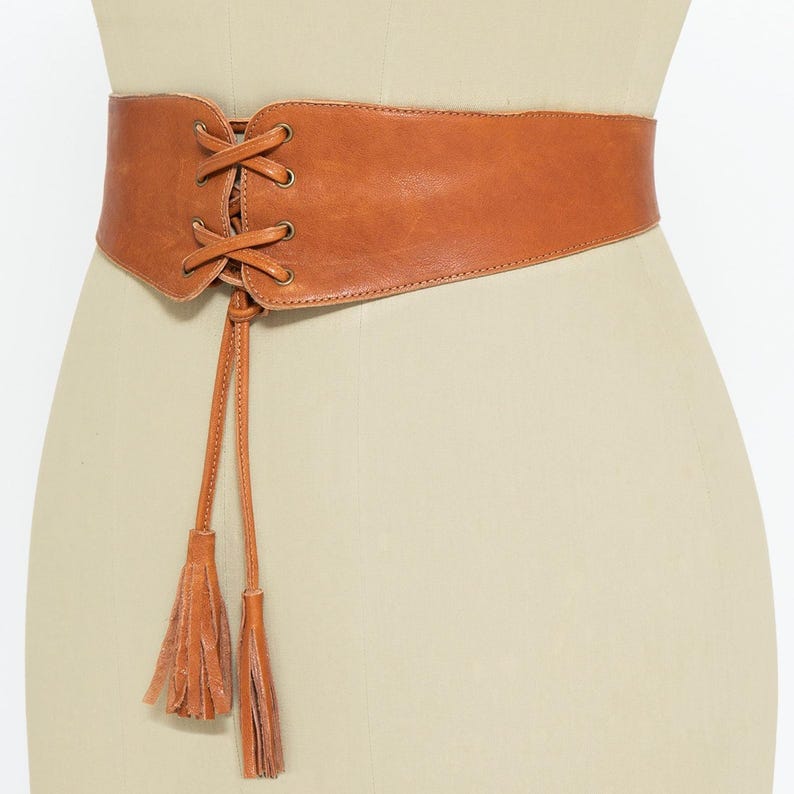 Back view of a handmade tan brown leather corset belt displayed on a mannequin. The belt is secured with three metallic snap closures, ensuring a comfortable and adjustable fit. The smooth, high-quality leather provides structure while allowing flexibility. At the front (not seen in the picture), it features a lace-up design with metal eyelets and long leather ties ending in tassels, enhancing its bohemian and waist-cinching style.