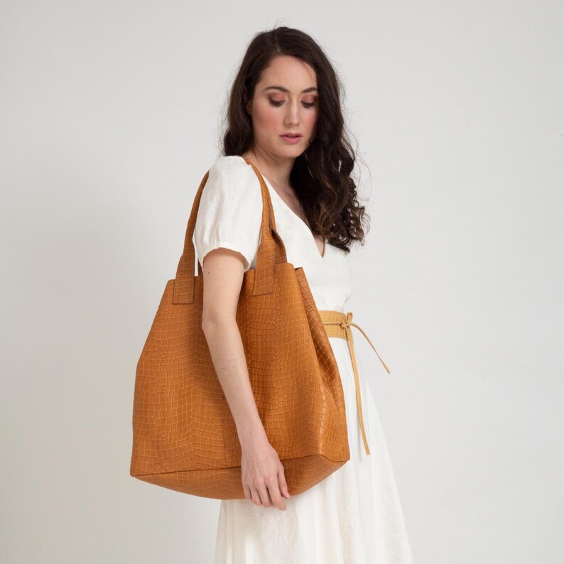 May include: A large, structured tote bag in a warm mustard-yellow hue featuring a textured, embossed crocodile-skin pattern. The bag has two sturdy shoulder straps and a spacious, open-top design, crafted with a consistent finish throughout.