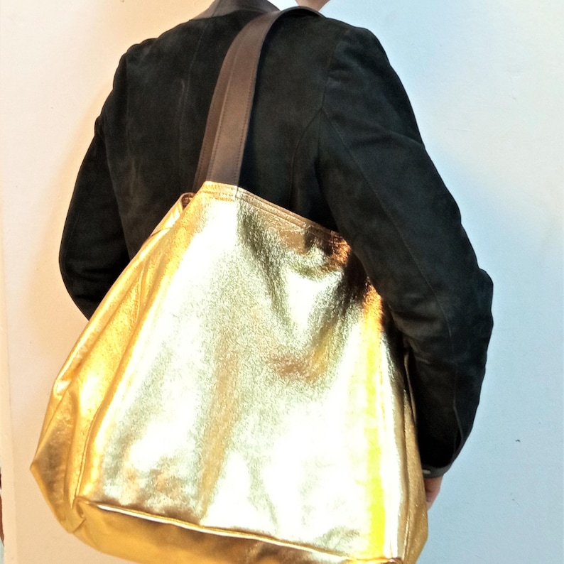 Puede incluir: Un bolso tote grande de color dorado con una correa de cuero marr&oacute;n. El bolso est&aacute; hecho de un material suave y texturizado y tiene un interior espacioso.