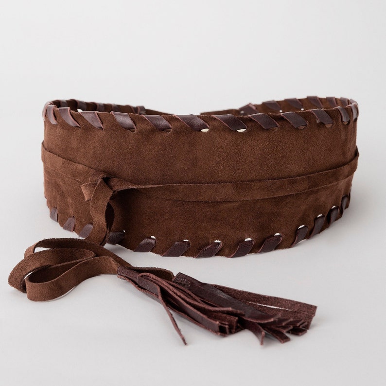A brown suede obi belt displayed against a light neutral background. The belt is coiled to showcase its wide design, decorative napa leather lacing along the edges, and long suede ties with tassel details. Handmade from soft chocolate-brown leather, this adjustable waist belt has a boho-chic style, perfect for pairing with dresses, tunics, and oversized shirts.