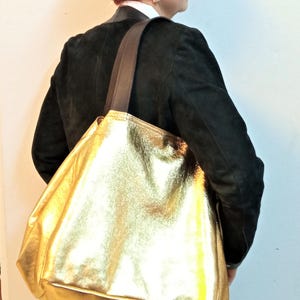 May include: A large gold leather tote bag with brown leather straps. The bag is being carried by a person wearing a black jacket.