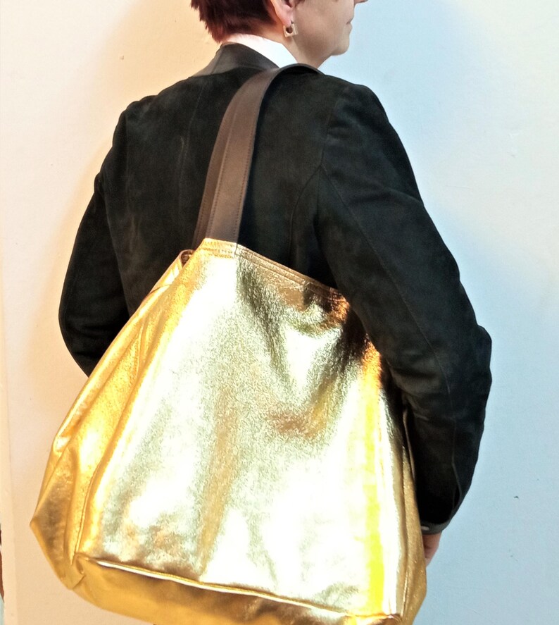 May include: A large gold leather tote bag with brown leather straps. The bag is being carried by a person wearing a black jacket.