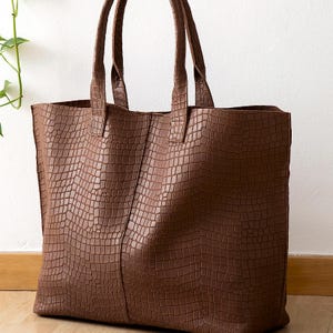 May include: A brown tote bag featuring a textured crocodile skin pattern. The bag has two vertical stitched panels on the front and two matching brown shoulder straps attached to the top edge. The design is structured with a rectangular shape.