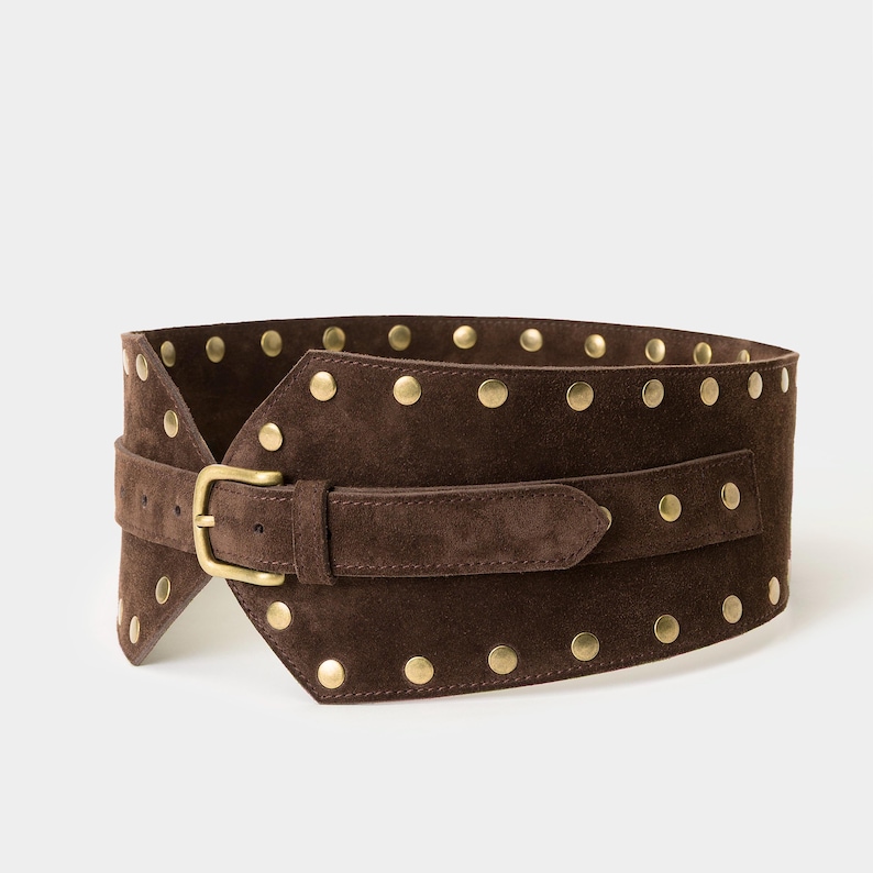 May include: A wide dark brown suede belt featuring a series of round brass-toned metal studs along the top and bottom edges. The belt includes a matching suede strap with a brass-toned metal buckle and a single loop for securing the end.