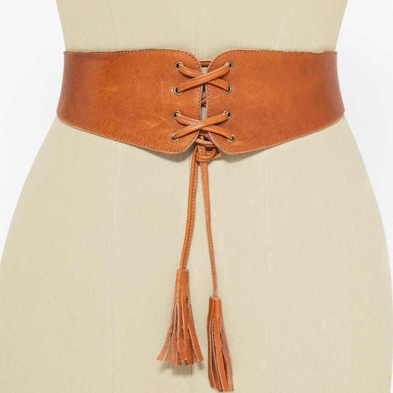 Front view of a handmade tan brown leather corset belt displayed on a mannequin. This waist-cinching belt features a lace-up front with metal eyelets and long leather ties finished with tassels. The curved design enhances the silhouette, while the back is adjusted with three clasp closures for a secure and comfortable fit. A stylish accessory perfect for dresses, tunics, blazers, and oversized shirts