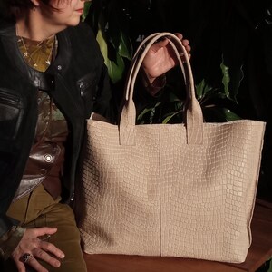 May include: A large tote bag in a light beige color featuring a textured, embossed grid pattern that resembles reptile skin. The bag has two sturdy top handles and a rectangular shape with a vertical seam running down the center of the front panel.