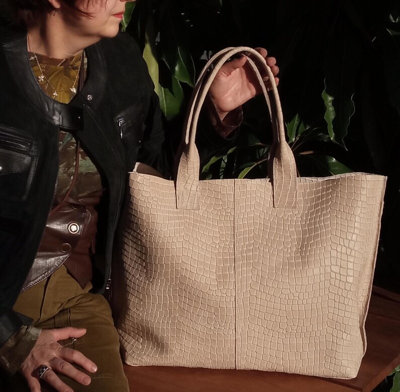 May include: A large tote bag in a light beige color featuring a textured, embossed grid pattern that resembles reptile skin. The bag has two sturdy top handles and a rectangular shape with a vertical seam running down the center of the front panel.